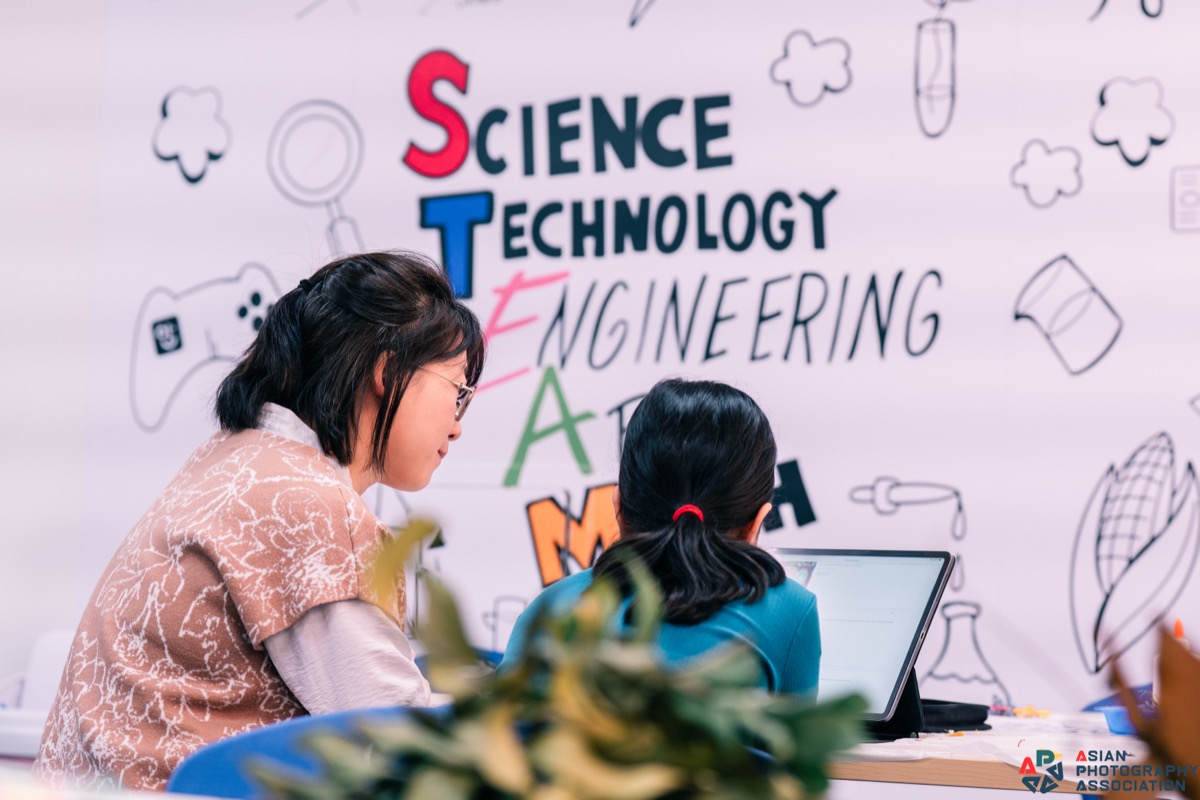 A KindleWood facilitator sitting beside a child at a laptop, in front of a STEAM-themed studio wall.
